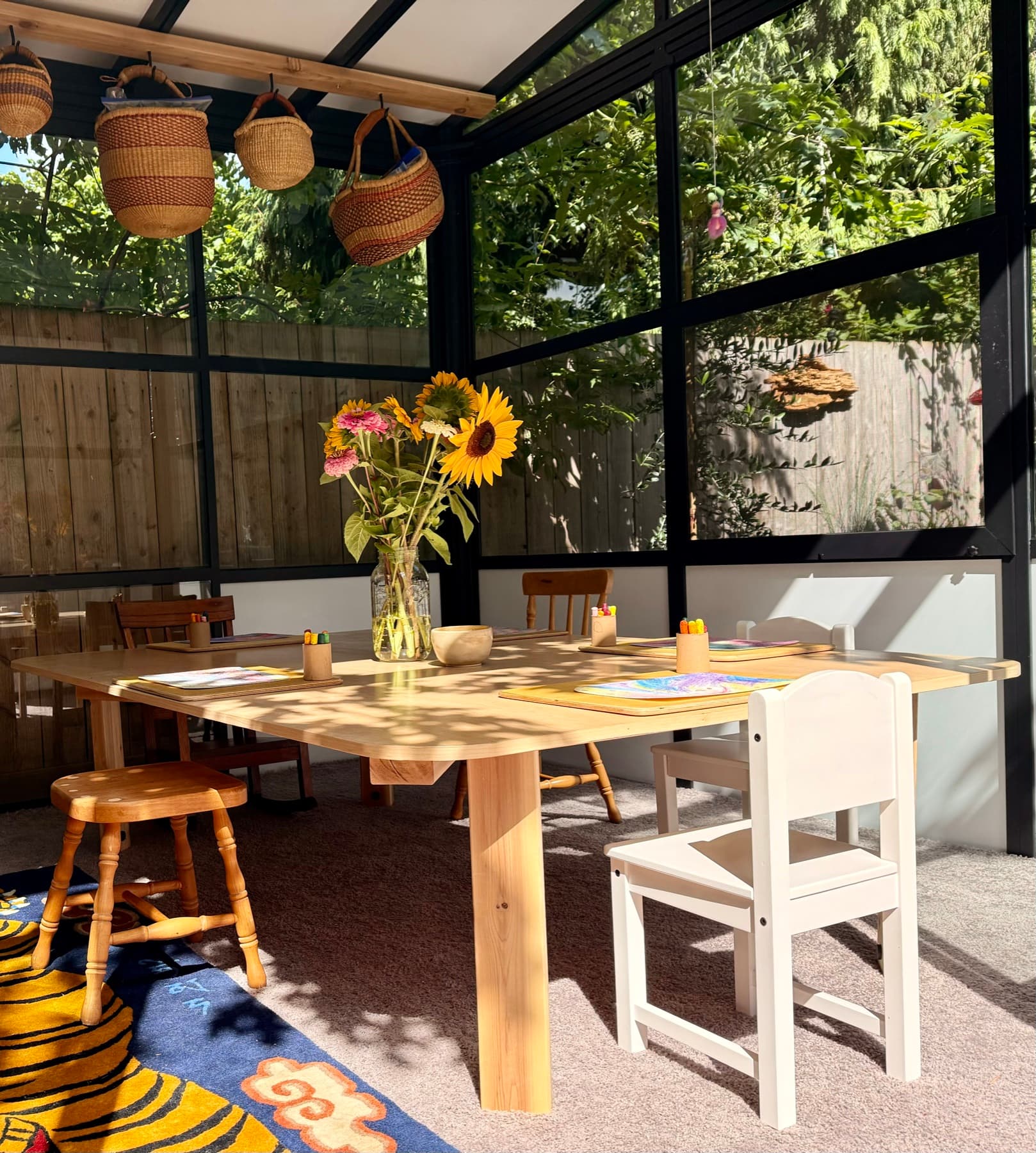 A sunlit studio table set with crayon drawings, sunflowers, and baskets overhead